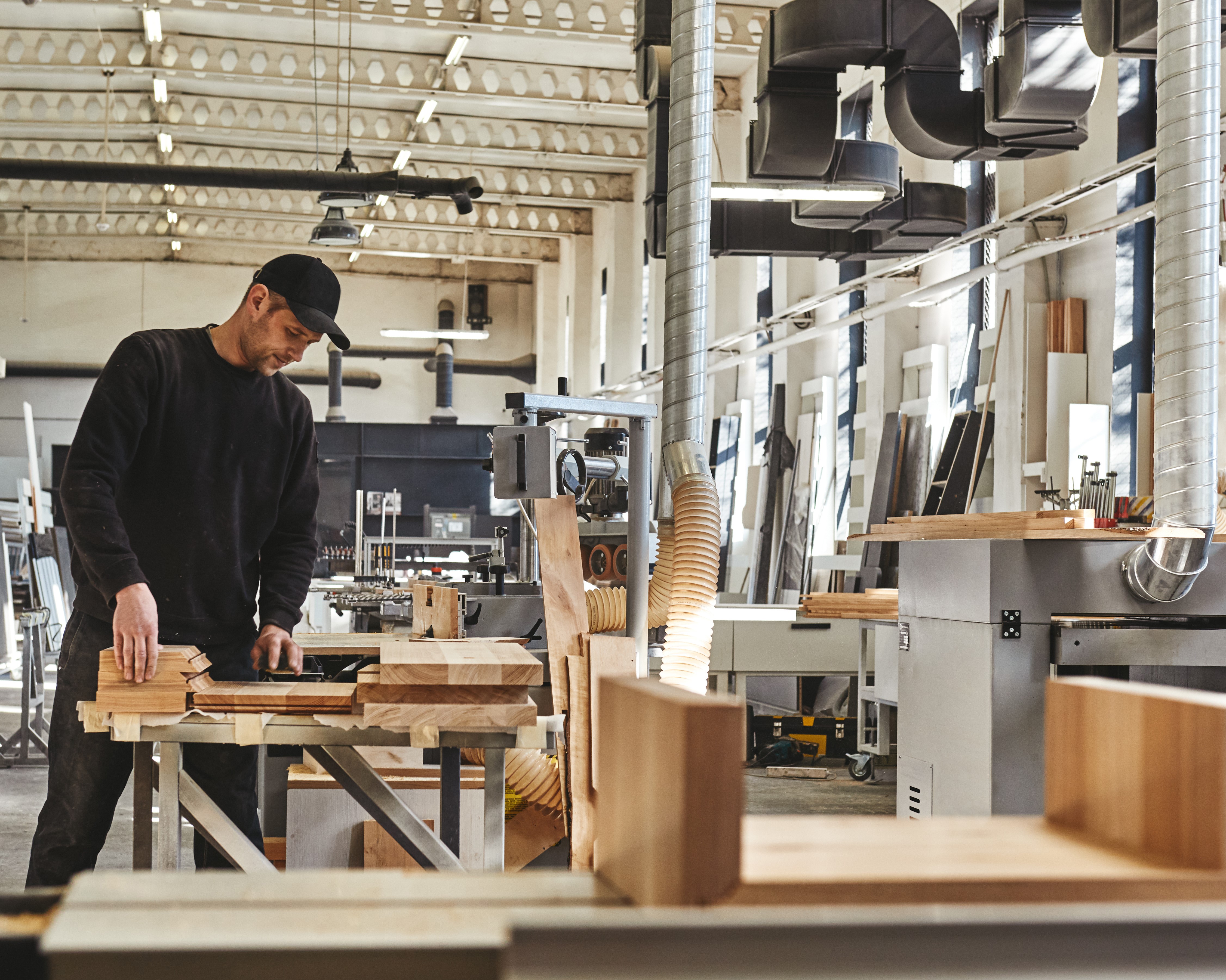 A man in a black cap and sweater works with wooden planks in a well-lit workshop with various carpentry tools and machines.